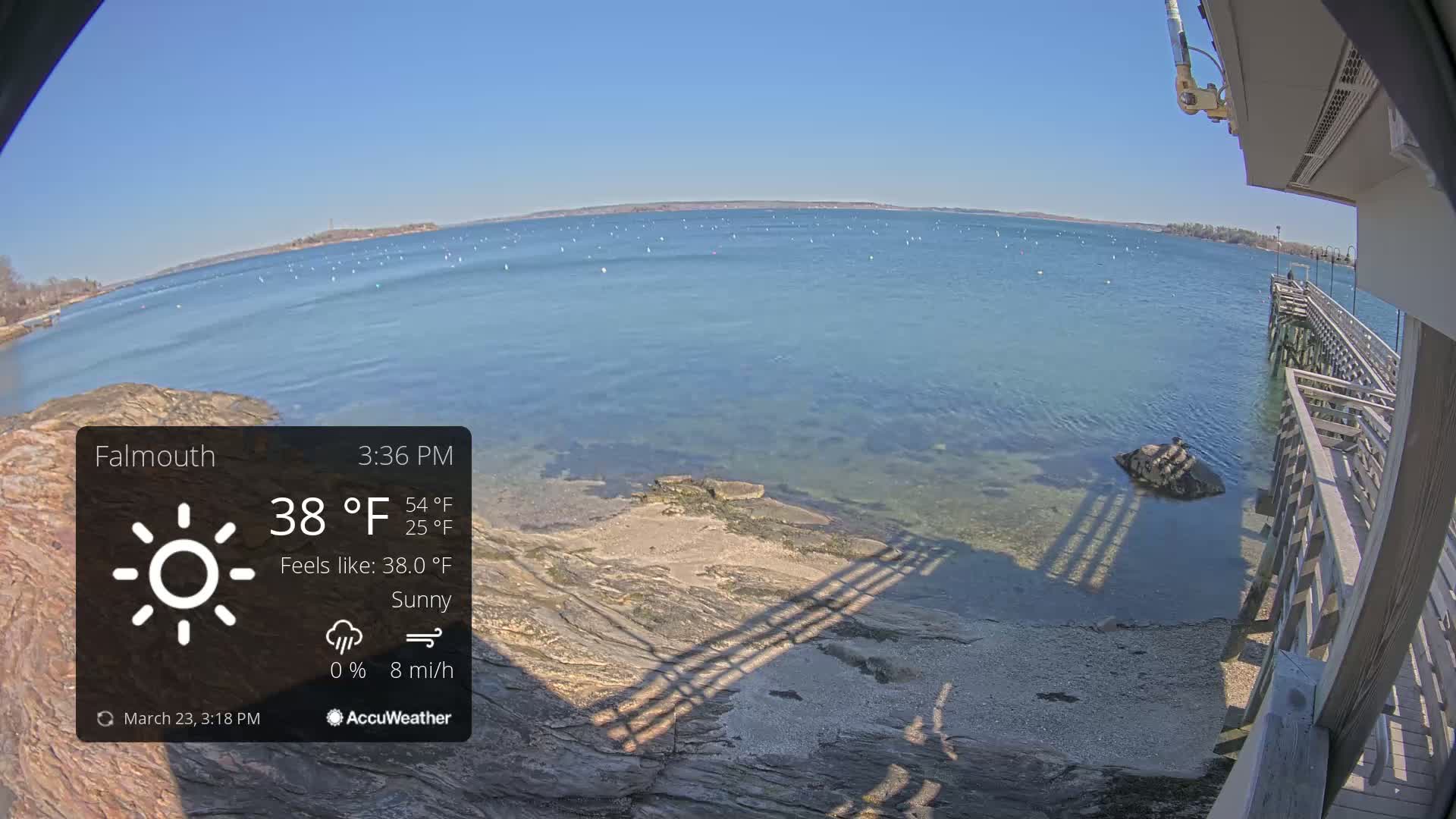 A sunny waterfront scene shows calm blue water, a rocky shoreline, and a wooden pier, with a slight breeze.