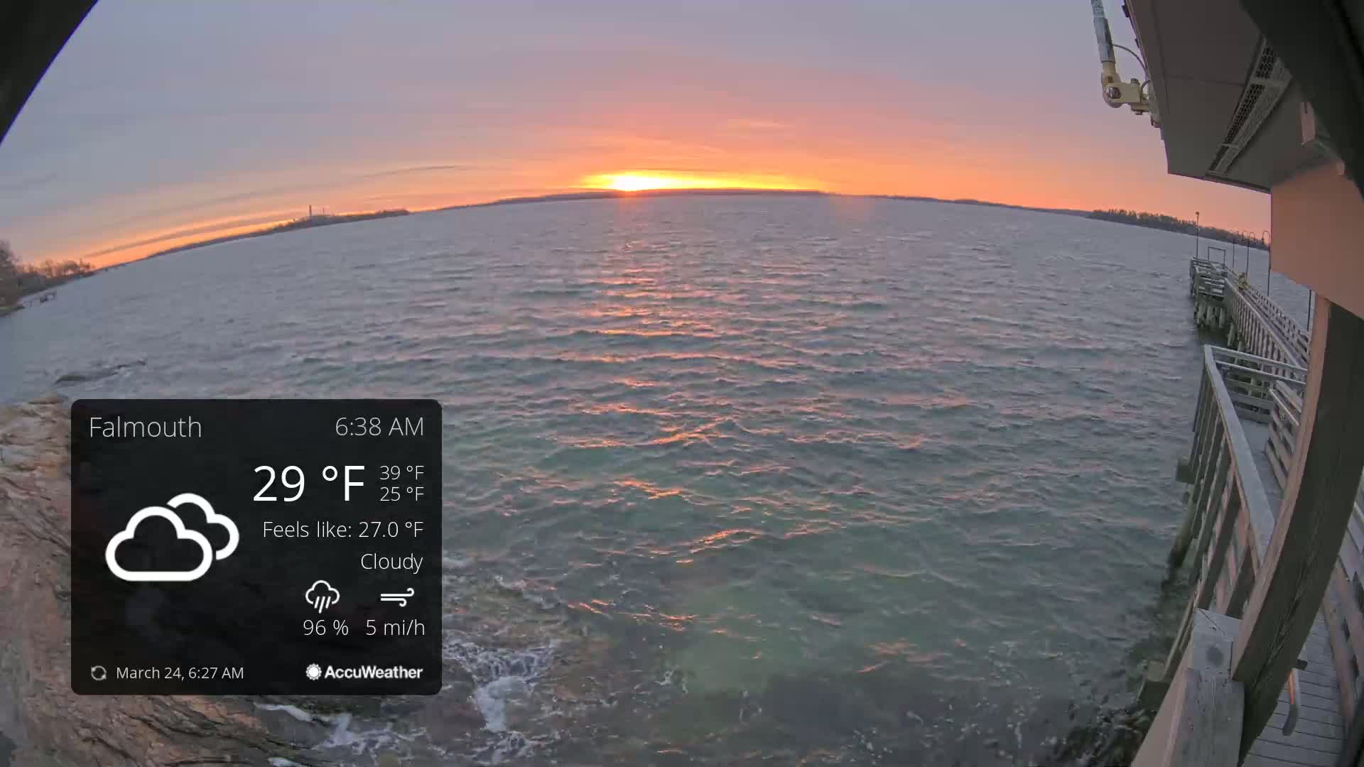 A cloudy sunrise casts an orange glow over a calm body of water, partially visible from a wooden pier.