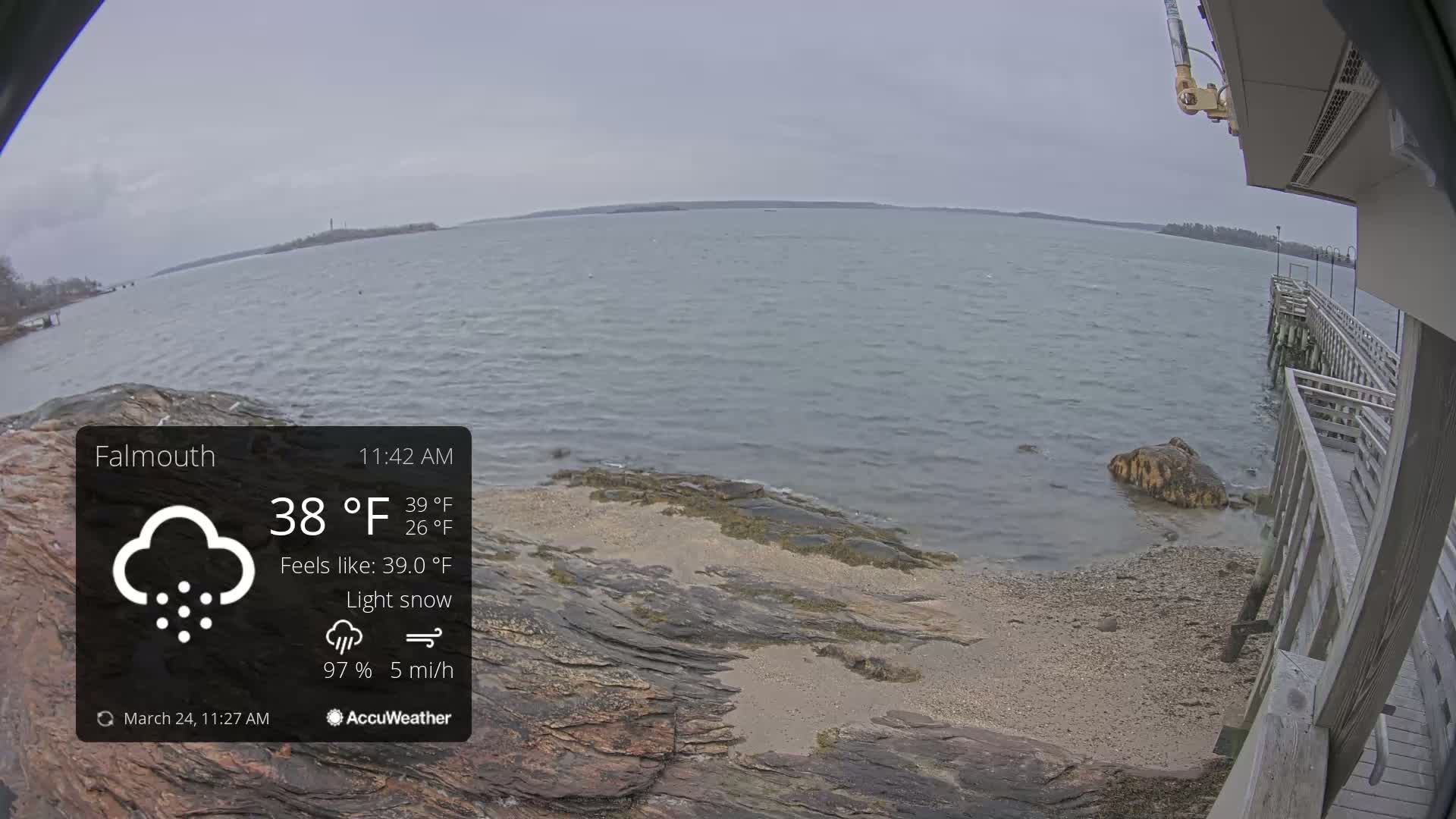 A rocky shoreline with a partly visible pier is seen on a lightly snowing, overcast day, with a calm body of water extending to a distant landmass.