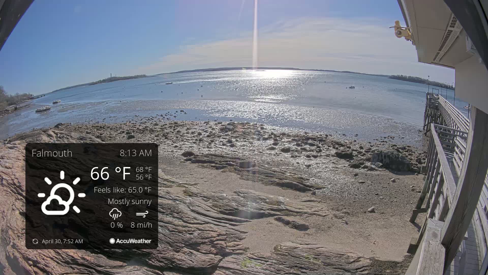 A mostly sunny, 66°F morning view of a rocky shoreline and calm bay with a wooden pier and distant landmass.