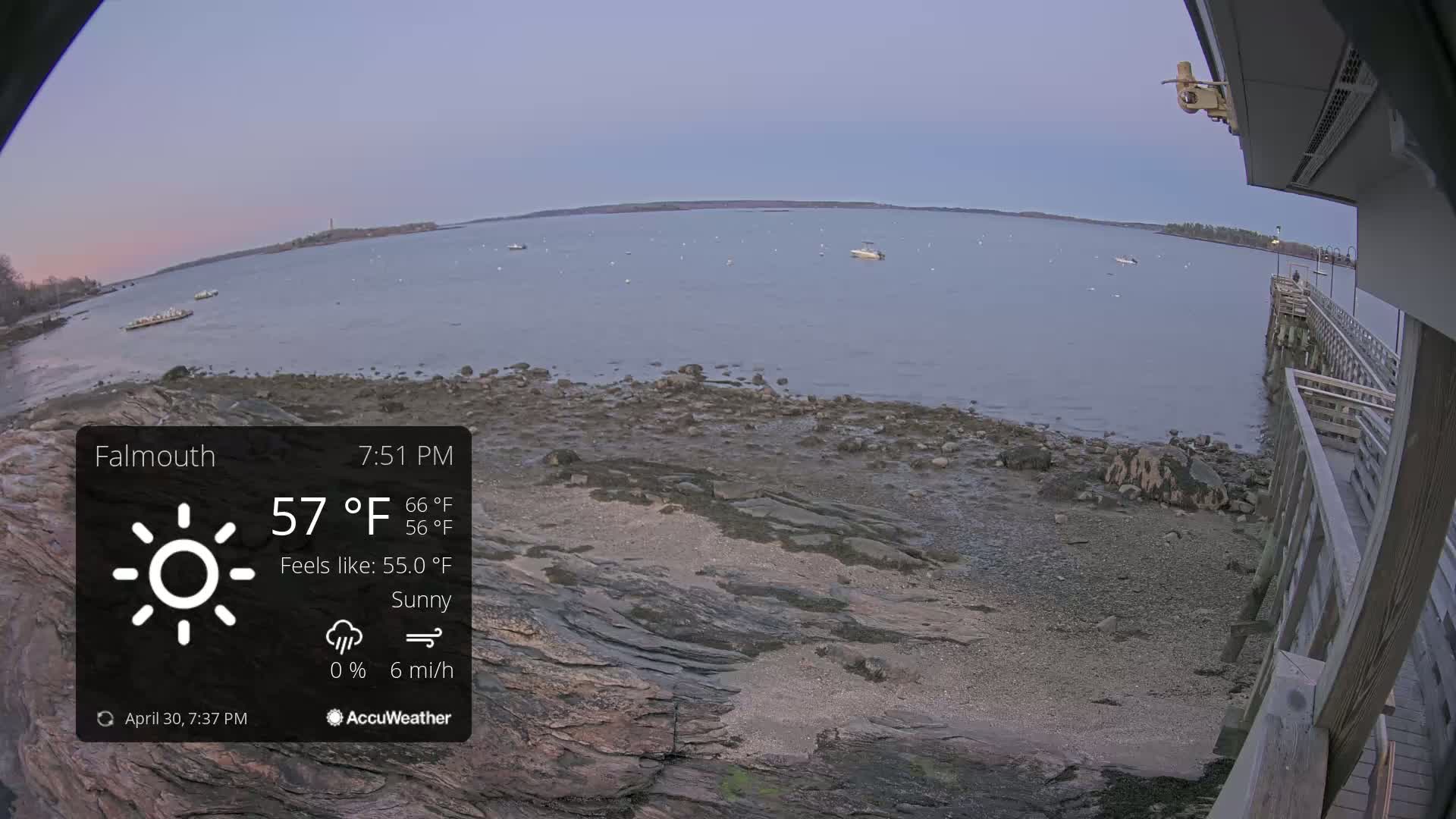 A sunny, calm day at a rocky shoreline with a wooden pier, boats on the water, and a distant landmass.