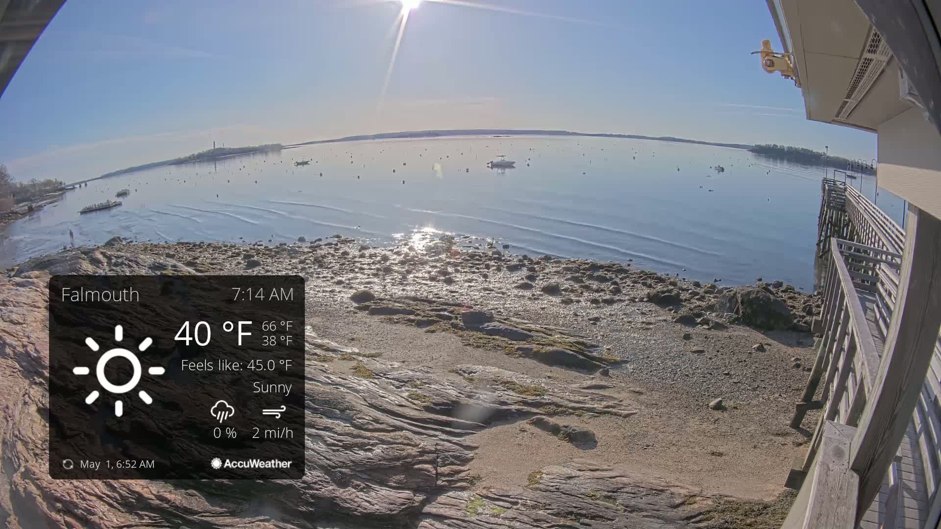 A sunny, calm morning scene shows a rocky shoreline with a wooden pier, a calm body of water with a few boats, and a distant landmass under a clear blue sky.