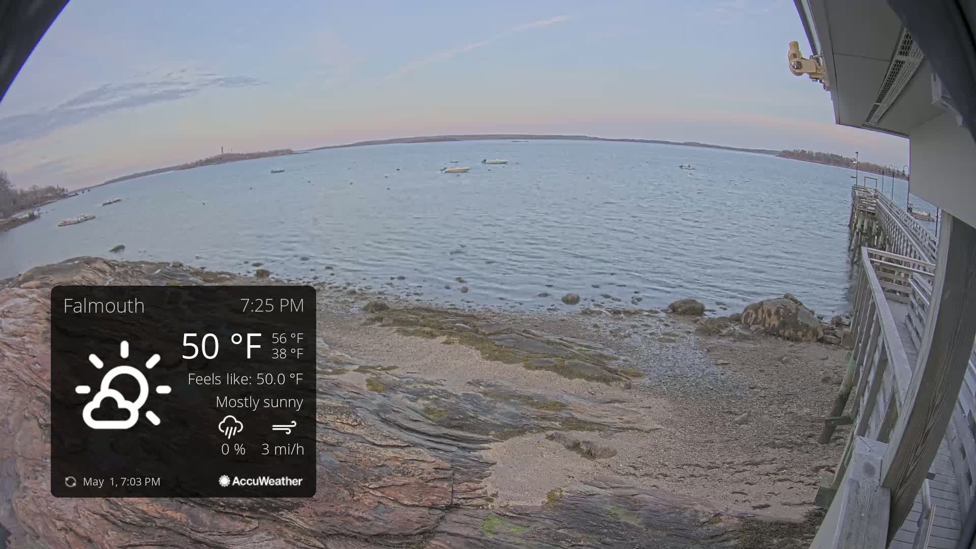 A mostly sunny, calm waterfront scene shows a rocky shoreline, a body of water with a few boats, and a wooden pier extending from a building.