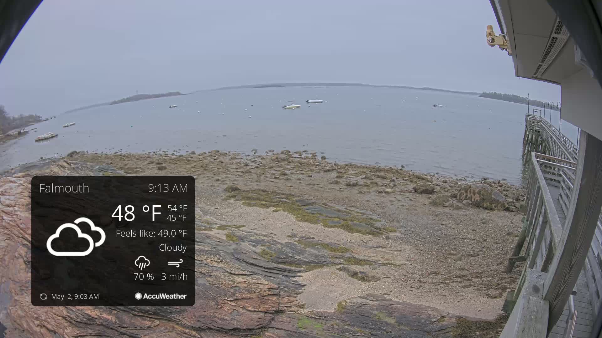 A cloudy, overcast day with a 3 mph wind shows a rocky shoreline and calm bay with several small boats; a wooden pier is visible in the foreground.
