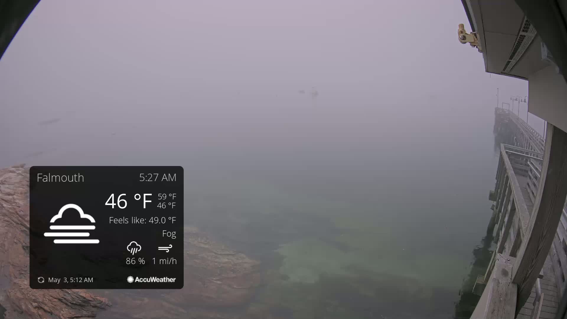 A foggy, low-visibility scene shows a wooden pier extending over calm water next to a rocky shoreline.
