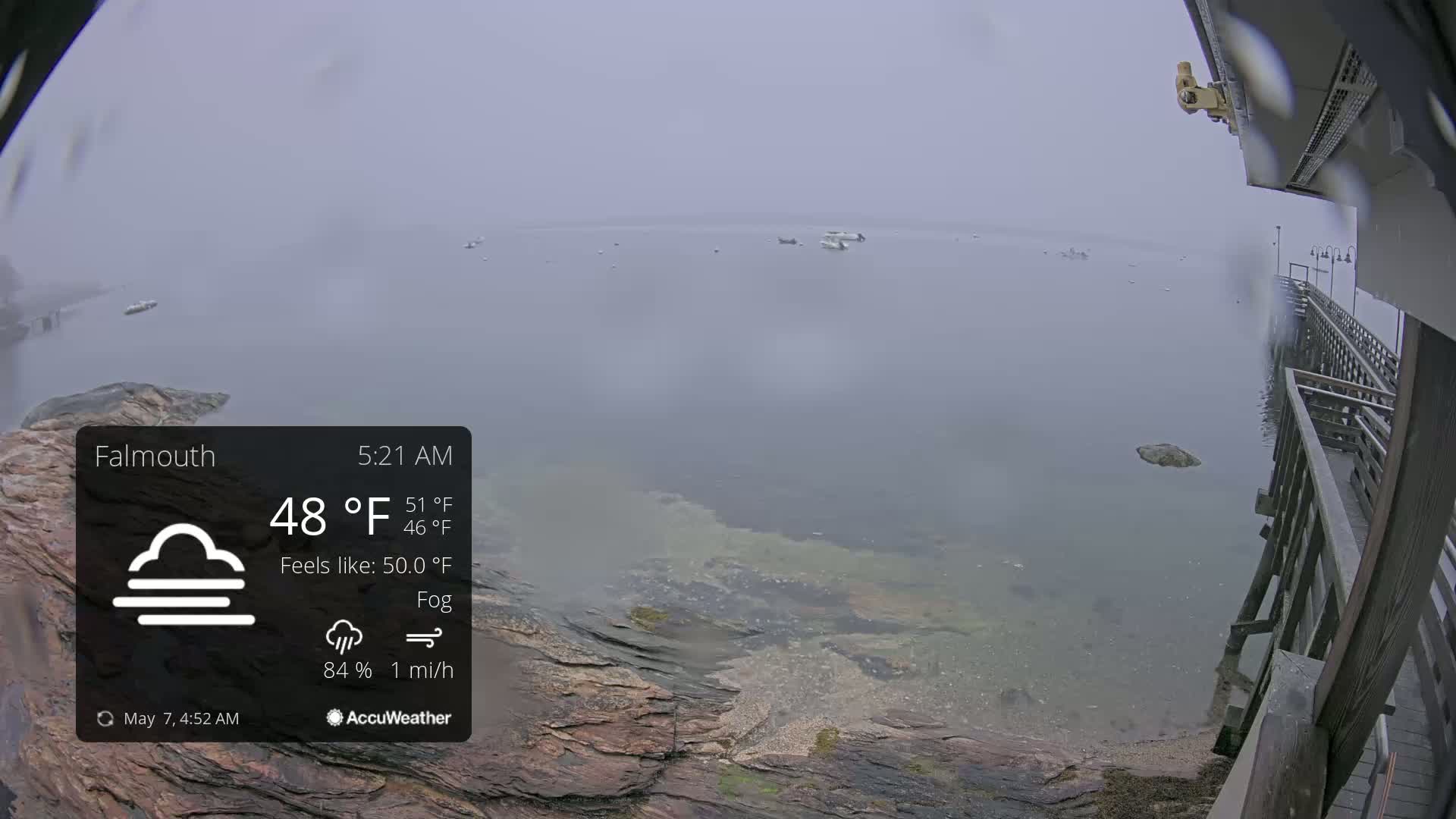 A foggy, calm body of water with several boats visible in the distance is seen from a wooden pier on a rocky shoreline.