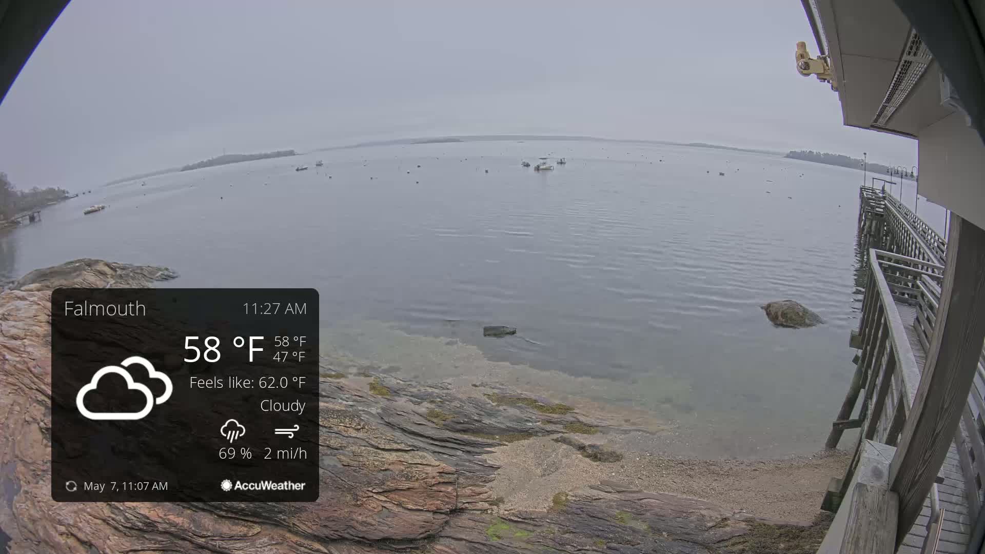 A cloudy, calm body of water with several boats and a rocky shoreline is visible from a wooden pier.