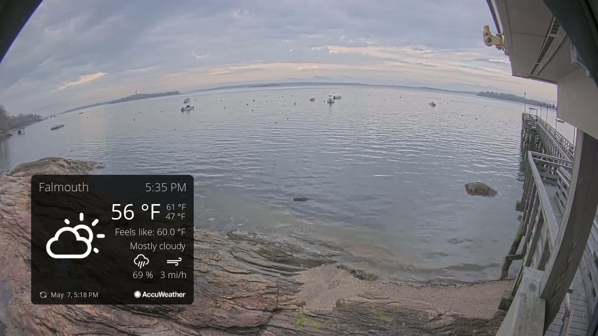 A mostly cloudy afternoon shows a calm body of water with several boats, a rocky shoreline, and a wooden pier extending from a building.
