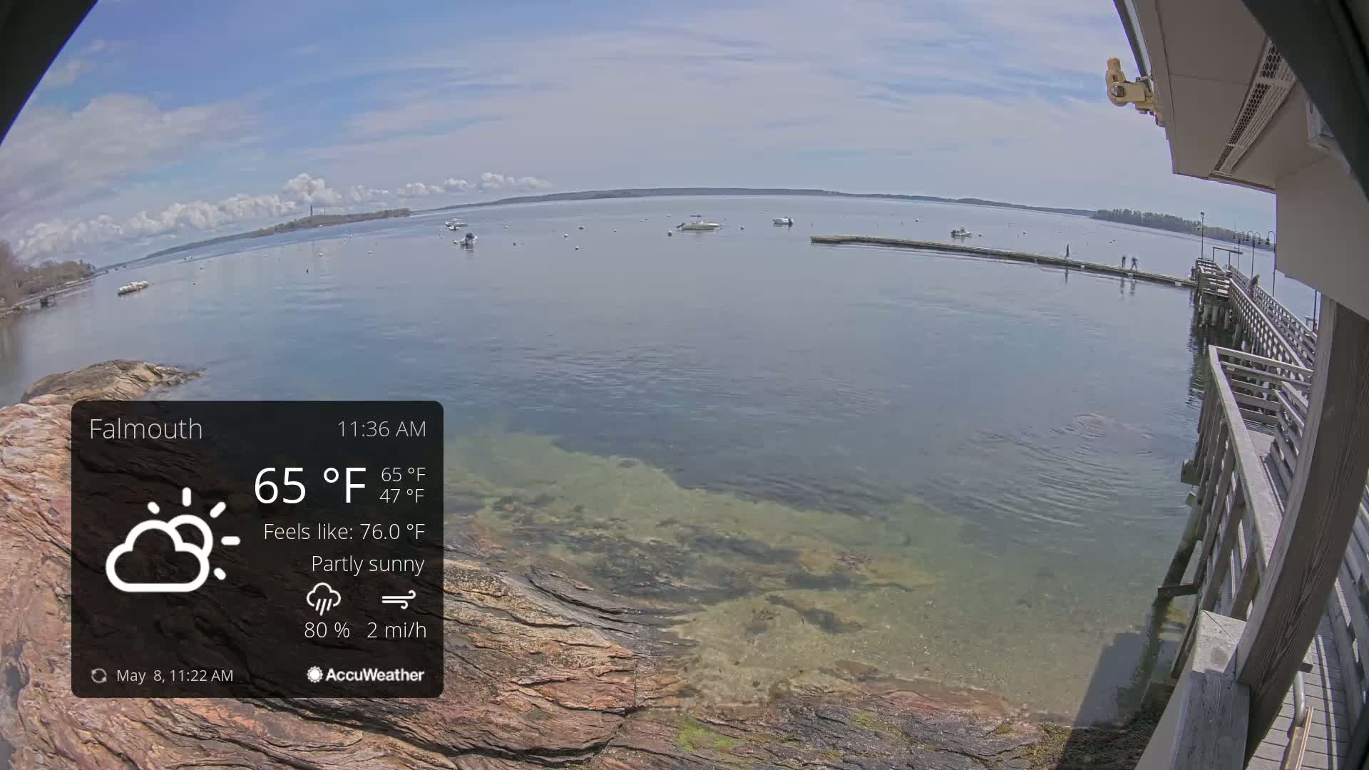 A partly sunny day shows a calm body of water with several boats and a wooden pier extending from a rocky shoreline.