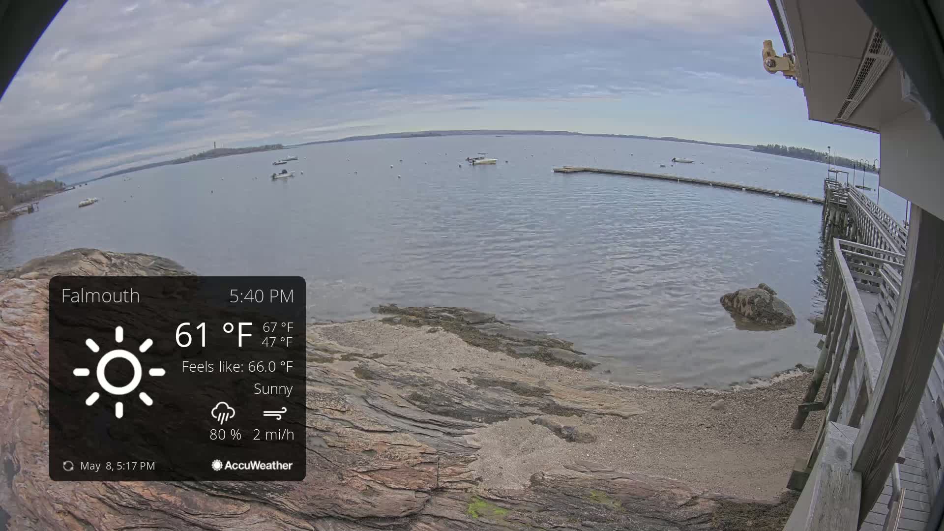 A sunny, breezy waterfront scene shows a rocky shoreline, calm water with several boats, and a wooden pier under a partly cloudy sky.