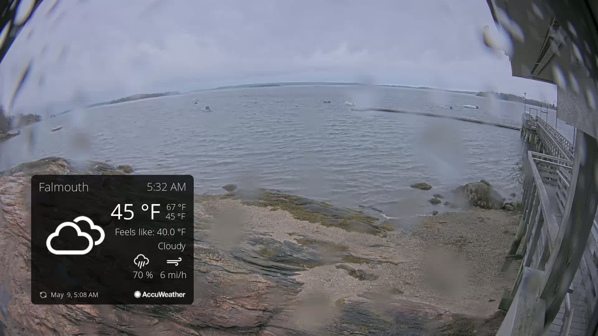 A cloudy, breezy morning scene shows a rocky shoreline with a wooden pier extending over a calm body of water, a few boats are visible in the distance.