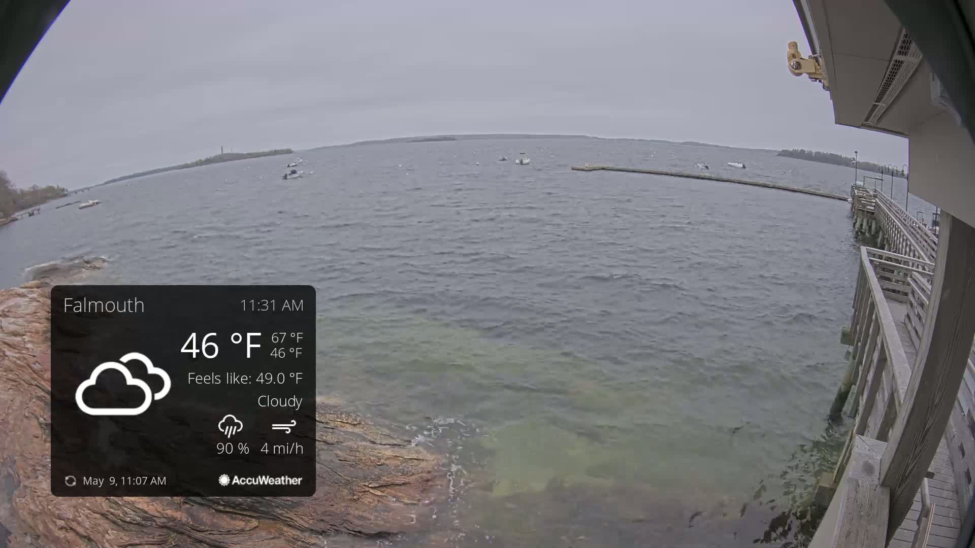 A cloudy, overcast day shows a body of water with a few boats, a rocky shore, and a wooden pier extending into the water.