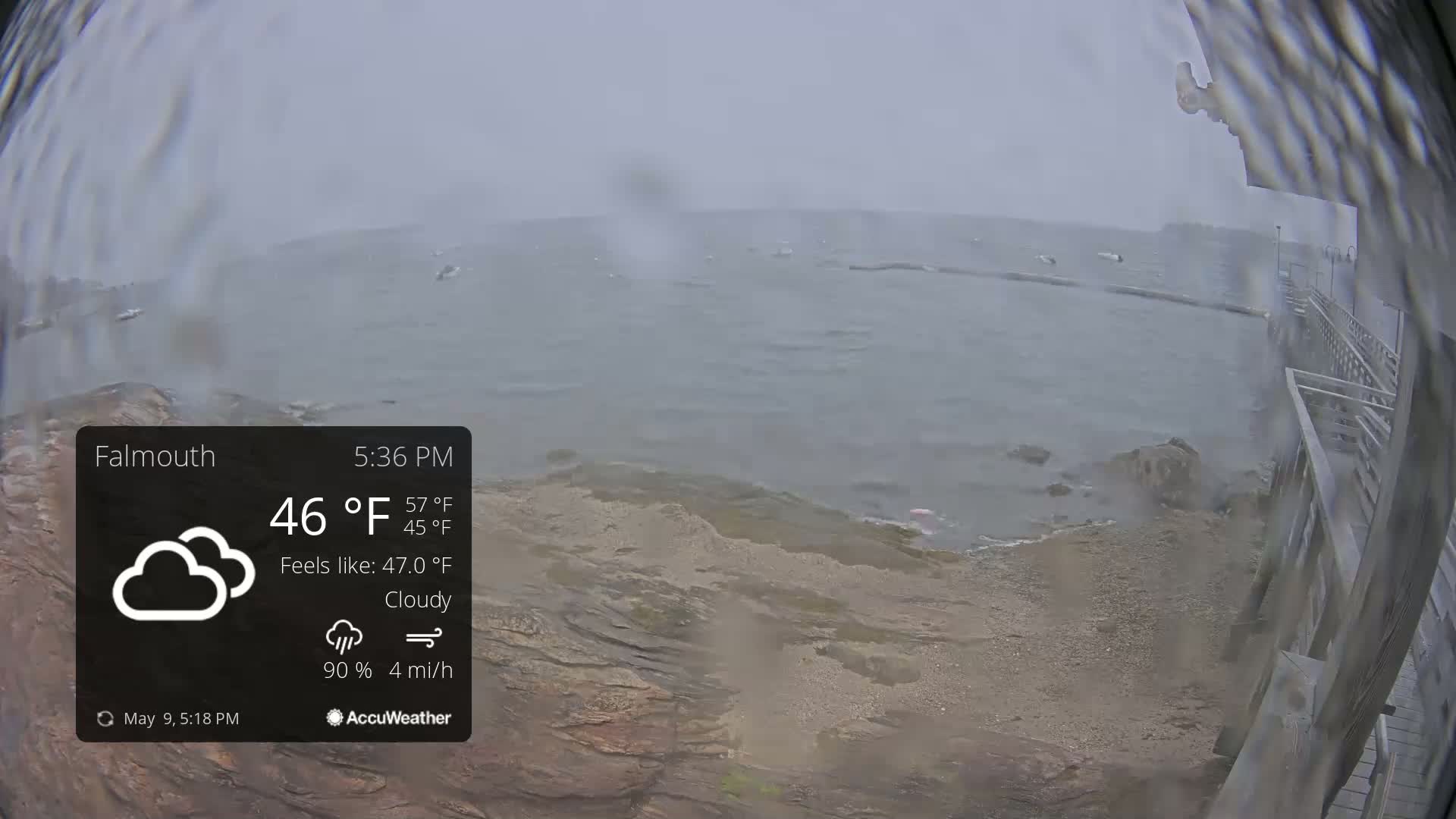 A cloudy, windy day with rain shows a rocky shoreline and calm ocean with a few boats in the distance, viewed from a partially visible structure.