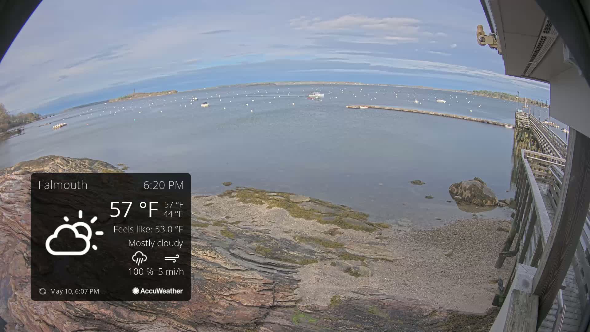 A mostly cloudy, 57°F day shows a rocky shoreline with a wooden pier and several boats on calm water.