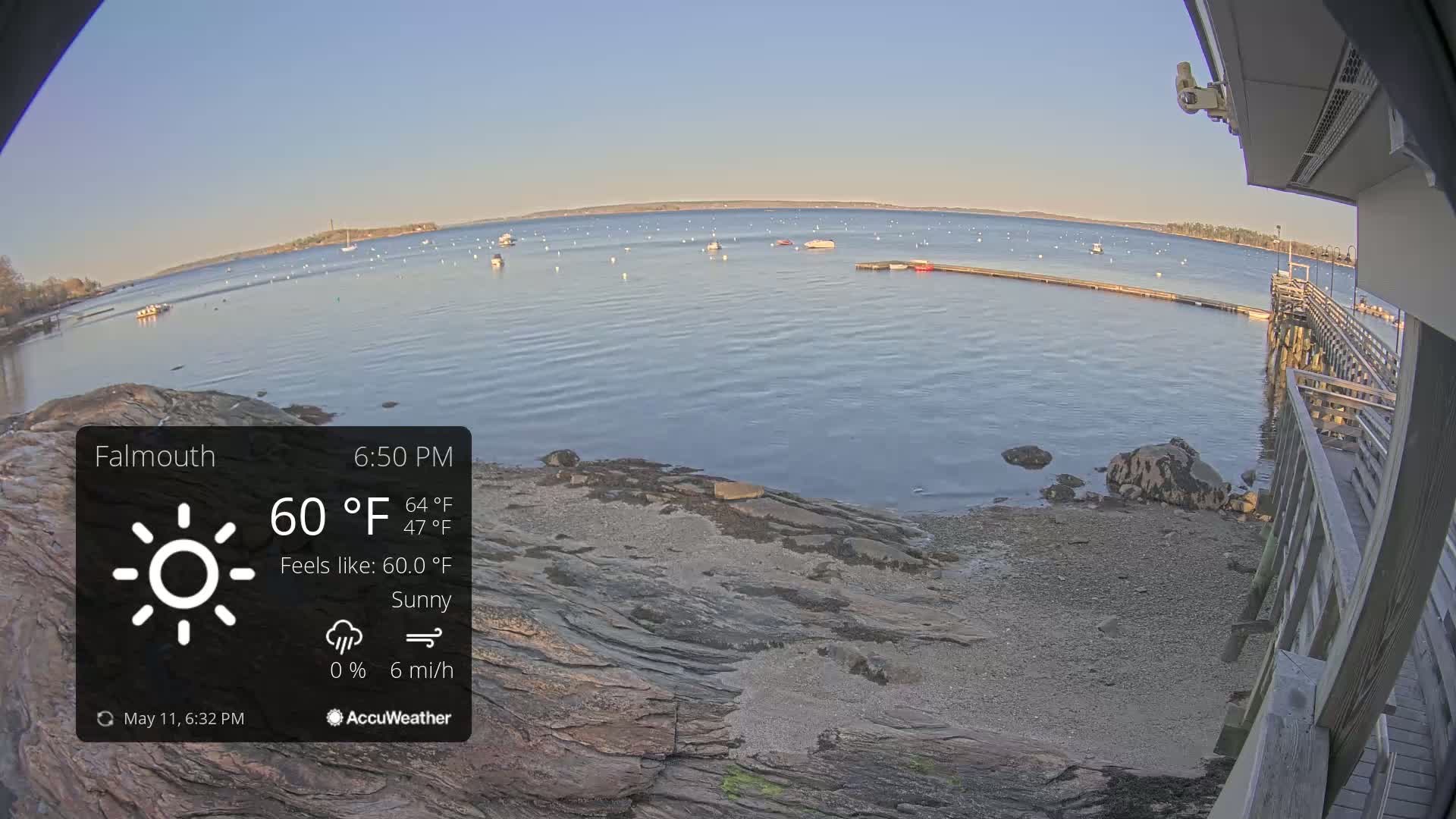 A sunny, calm bay with numerous boats is seen from a wooden pier on a rocky shoreline.