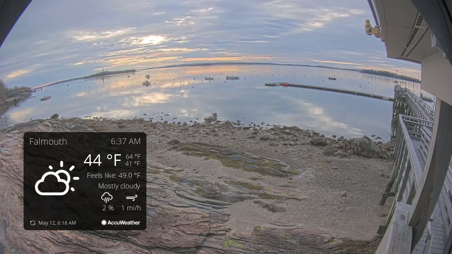 A mostly cloudy sunrise over a calm body of water with several boats, viewed from a rocky shoreline next to a wooden pier.