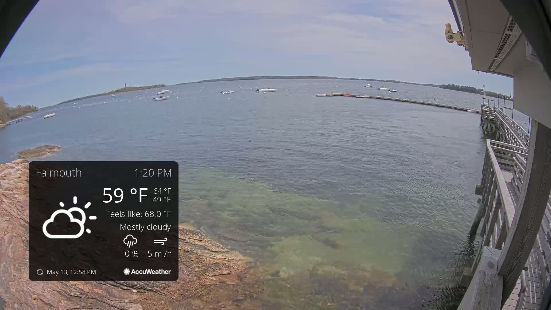 A mostly cloudy day with a 5 mph wind shows a view of calm water with several boats, a rocky shoreline, and a wooden pier.