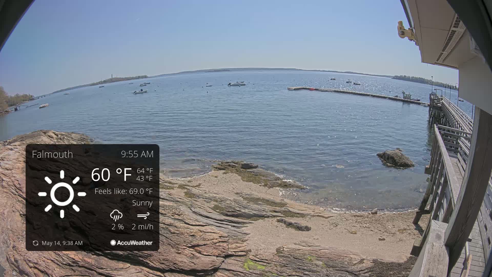 A sunny waterfront scene shows a calm body of water with several boats, a rocky shoreline, and a wooden pier extending from a building.