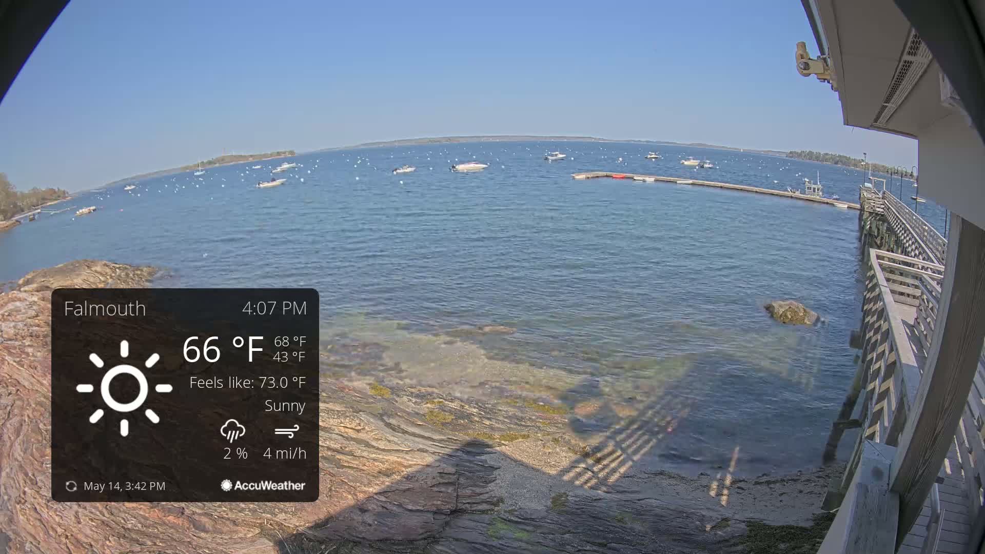 A sunny waterfront scene shows a calm body of water with several boats, a rocky shoreline, and a wooden pier on a bright day.