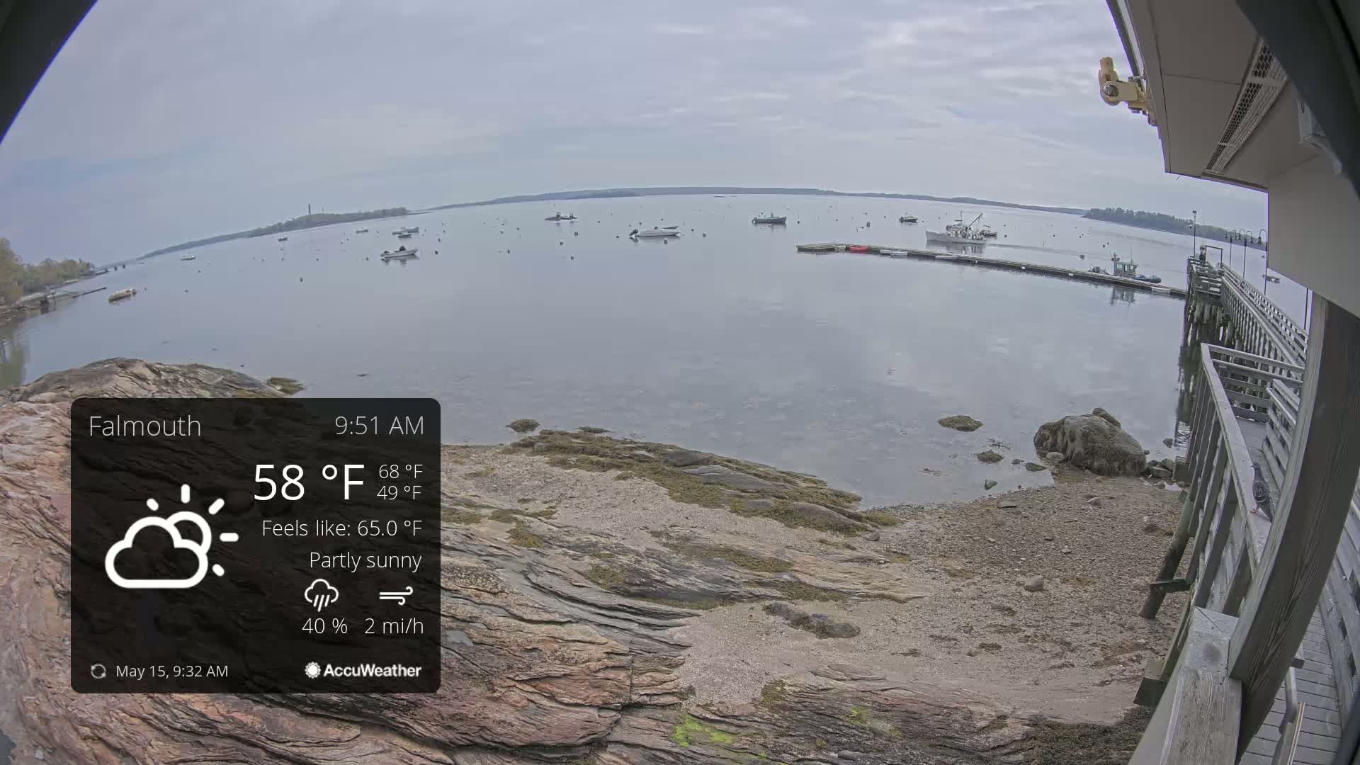 A partly sunny day with a 40% chance of rain and a 2 mph wind shows a calm body of water with several boats, a rocky shoreline, and a wooden pier.