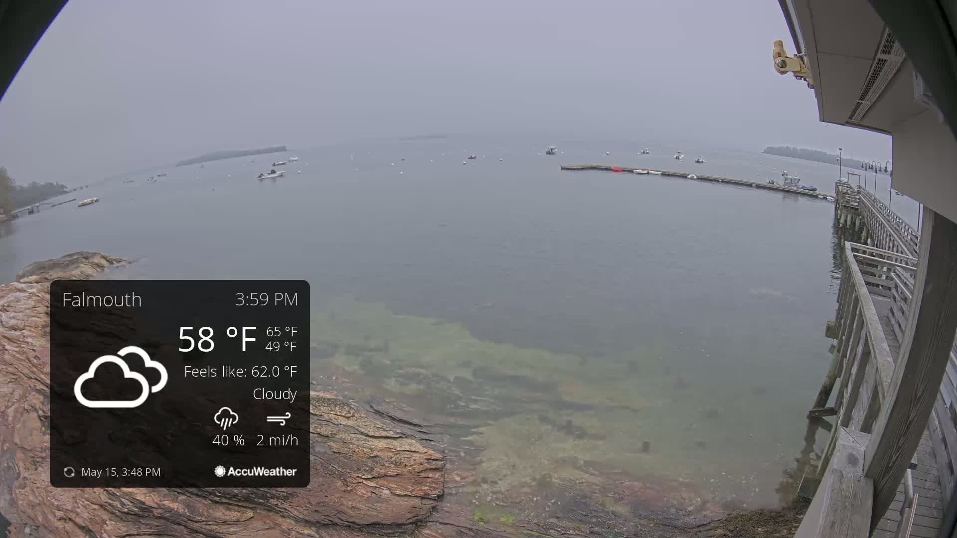 A cloudy, overcast day shows a view of a calm body of water with several boats and a wooden pier, partially visible from a rocky shoreline.