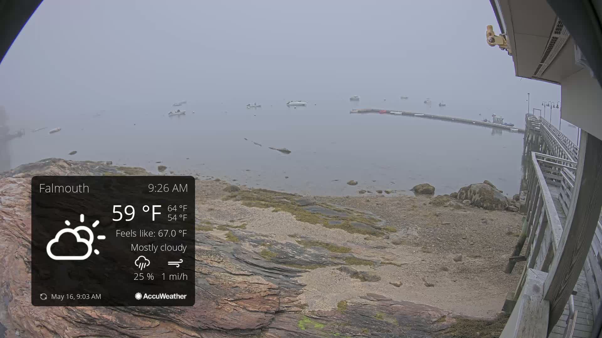 A foggy, mostly cloudy waterfront scene shows several boats in the distance, a rocky shore in the foreground, and a wooden pier to the right.