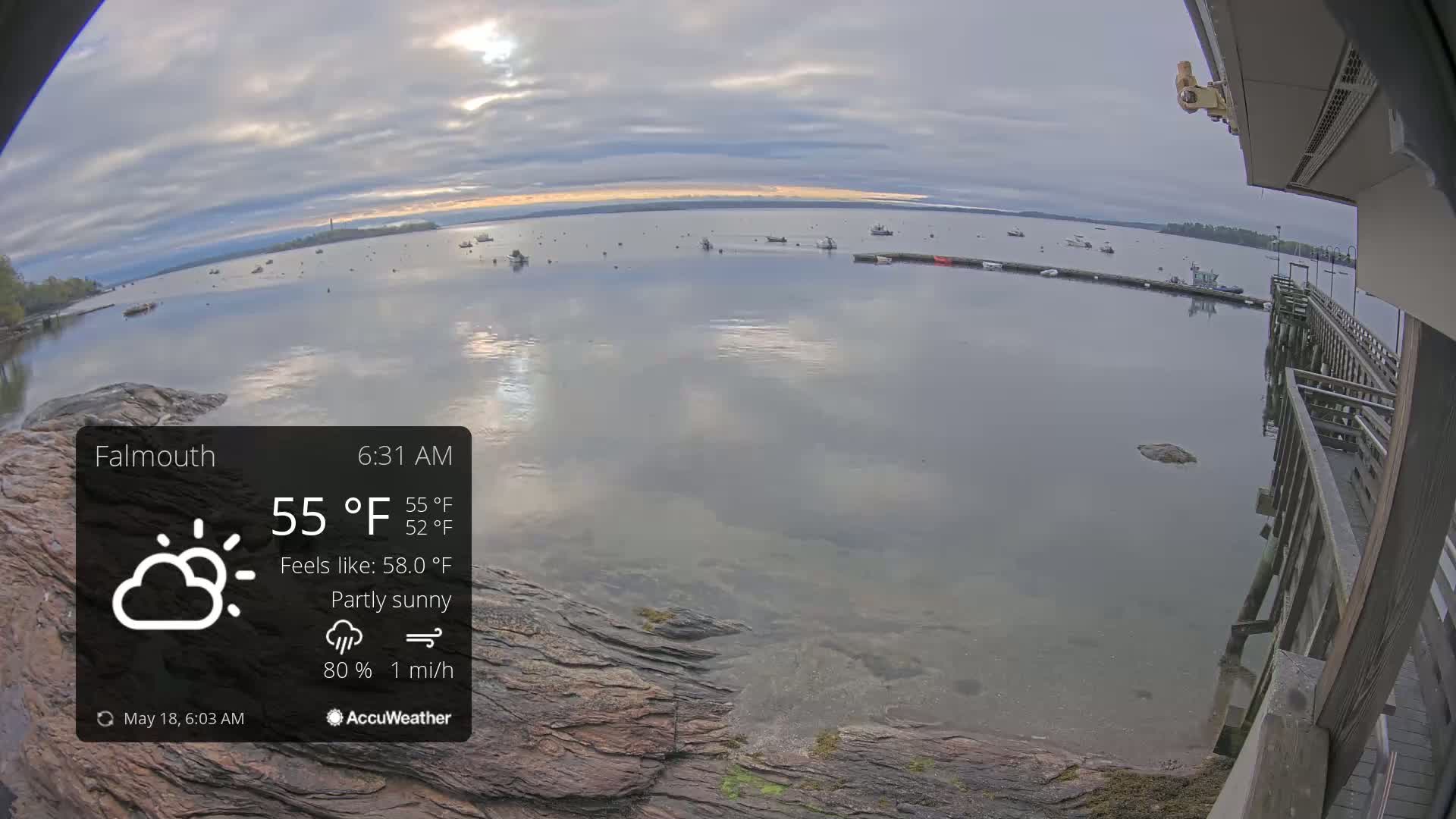 A partly sunny morning shows a calm body of water with several boats, viewed from a wooden pier on a rocky shore.