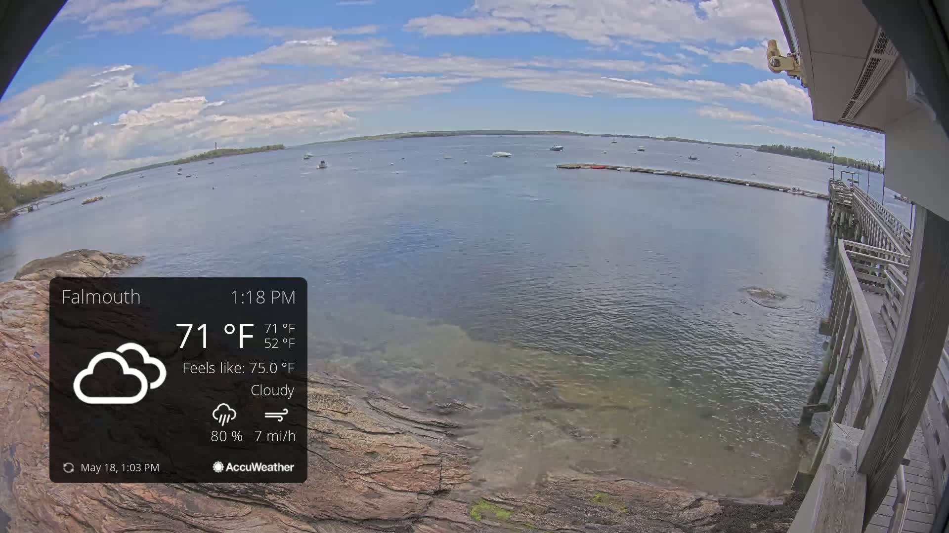 A partly cloudy day with a 7 mile per hour wind shows a calm body of water with several boats and a wooden pier extending from a rocky shoreline.
