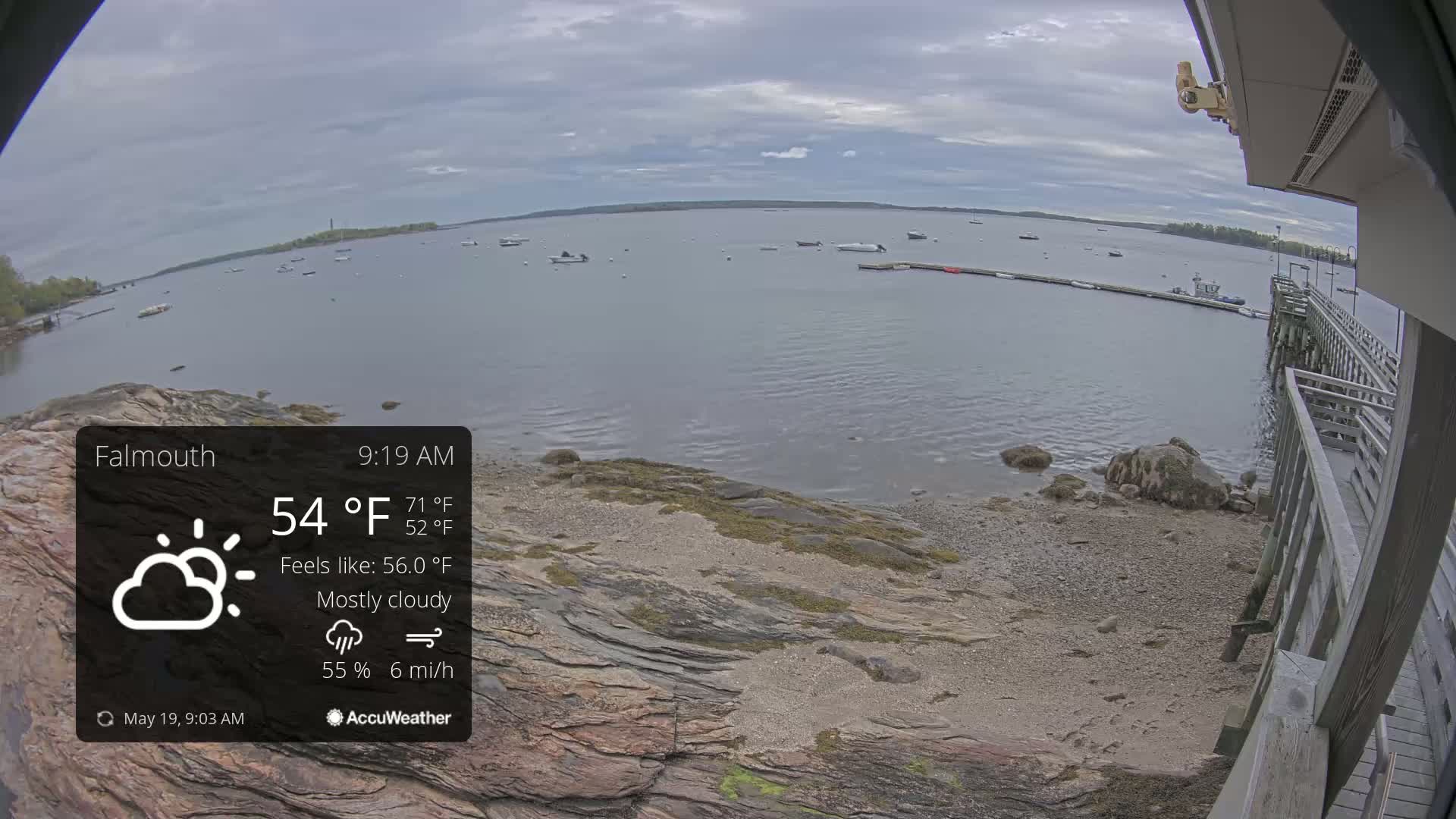 A mostly cloudy, 54°F day is shown overlooking a calm body of water with several boats, a rocky shoreline, and a wooden pier.
