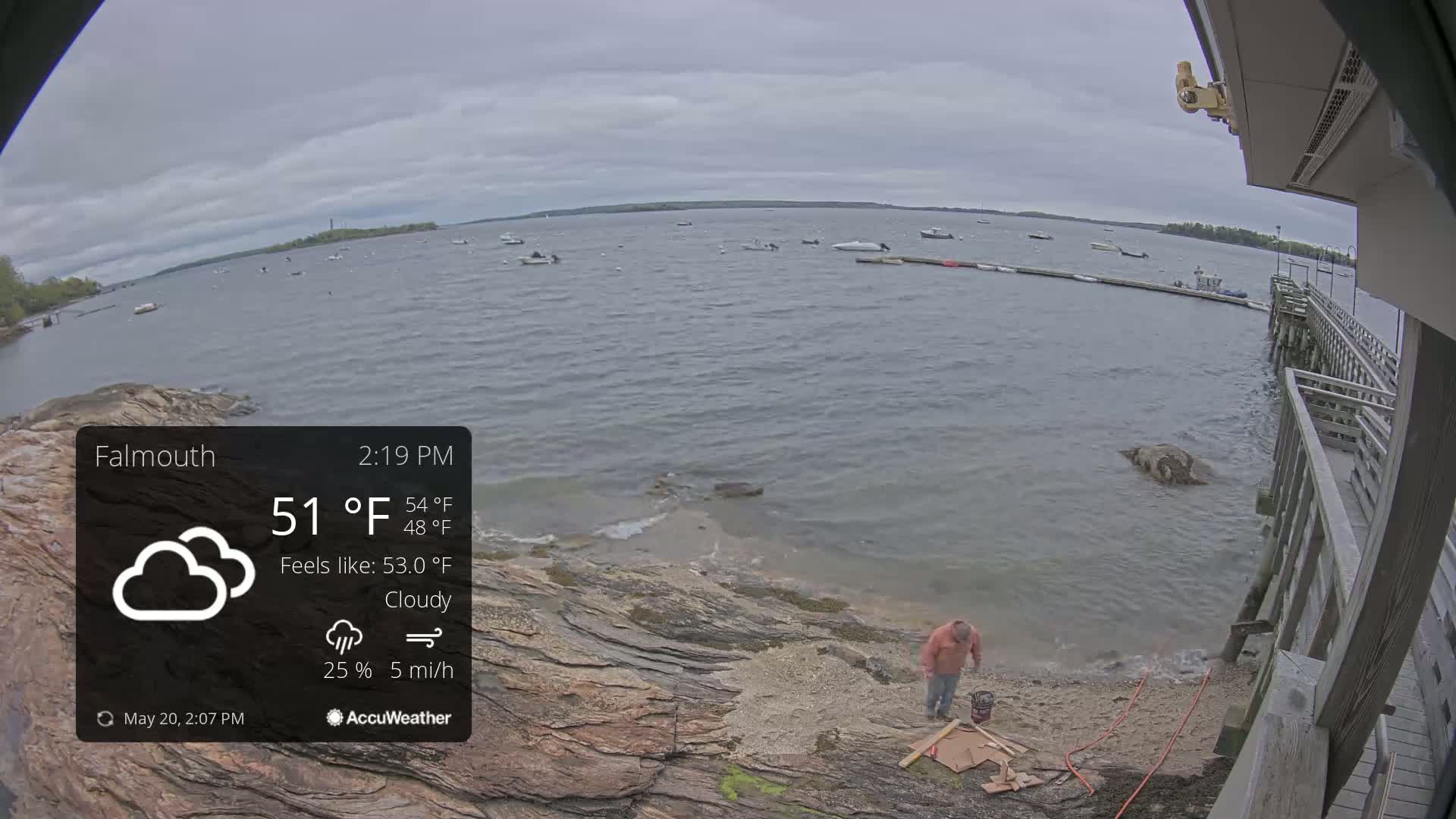 A person works near a rocky shoreline on a cloudy day, with several boats visible on the calm water in the background.