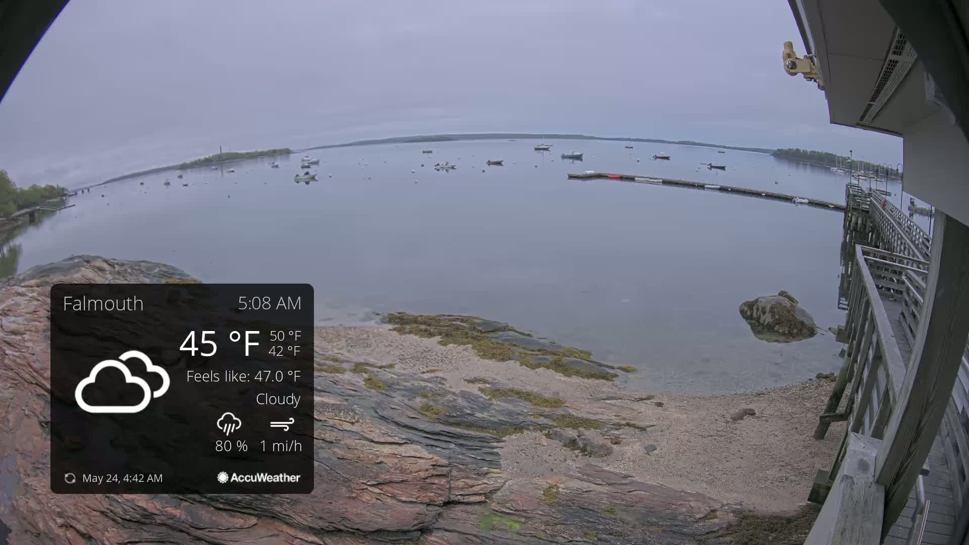 A cloudy, calm morning scene shows a rocky shoreline with a wooden pier overlooking a calm bay dotted with small boats.