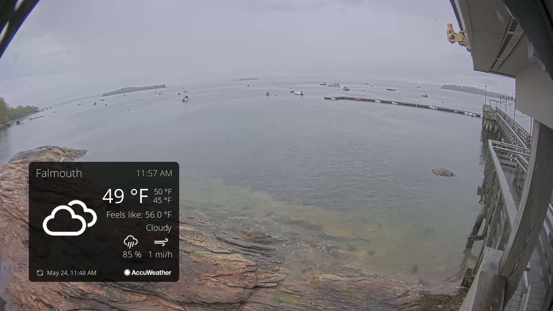 A cloudy, overcast day with a light breeze shows a calm body of water with several boats and a rocky shoreline; a wooden pier is visible in the foreground.