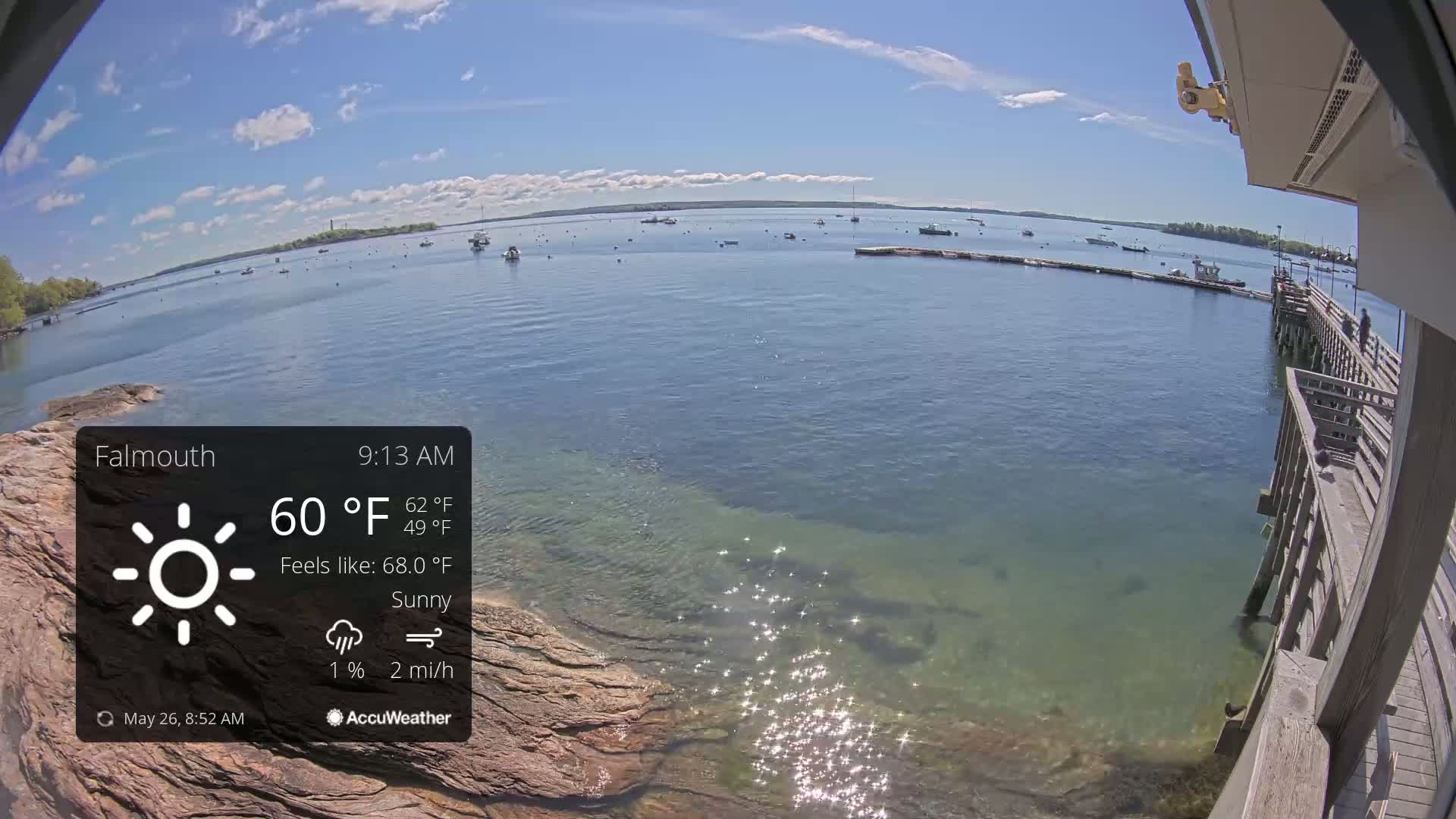 A sunny, calm day is shown overlooking a body of water with several boats, a rocky shoreline, and a wooden pier.