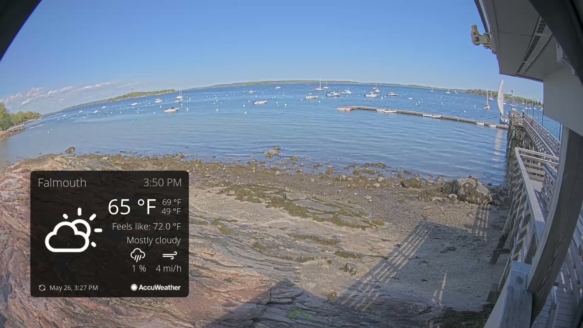 A mostly cloudy, calm day with a temperature of 65°F shows a view of a rocky shoreline, a body of water with numerous boats, and a wooden pier.