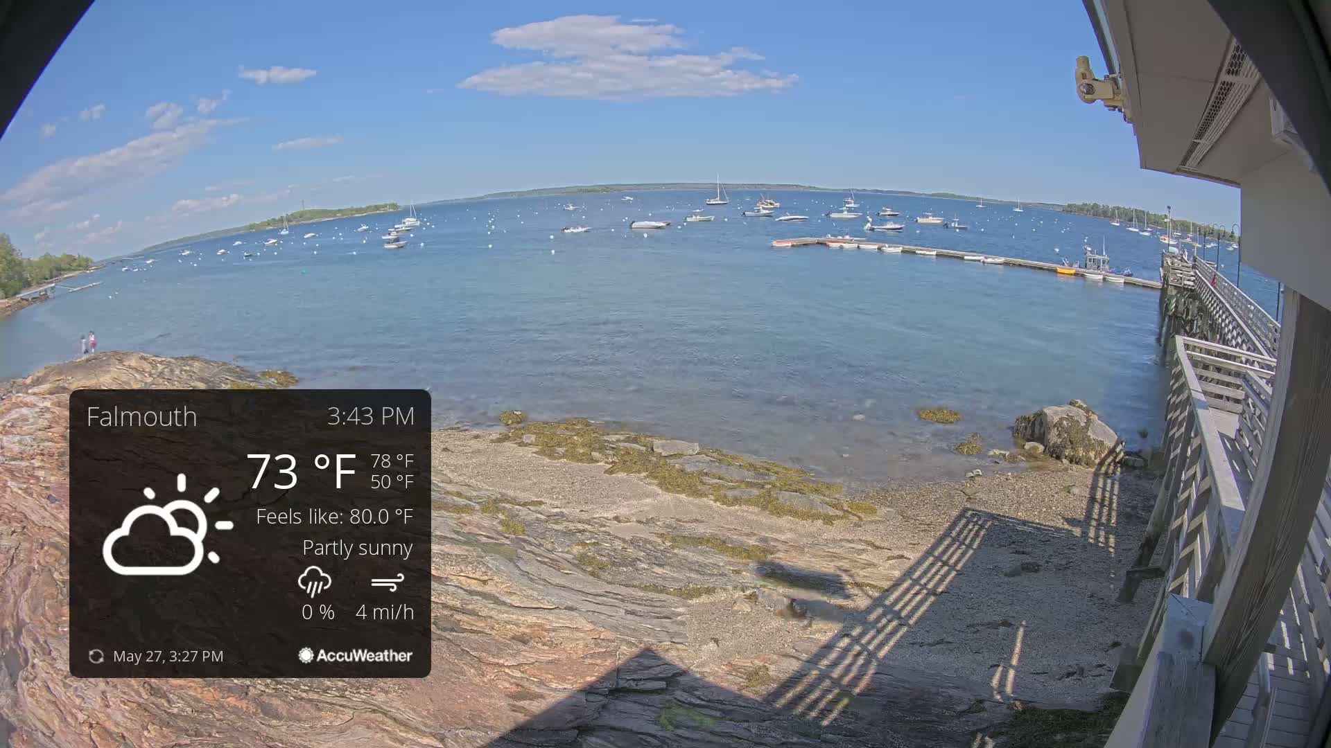 A partly sunny view of a calm body of water with numerous boats anchored in the distance, a rocky shoreline, and a wooden pier extending into the water.
