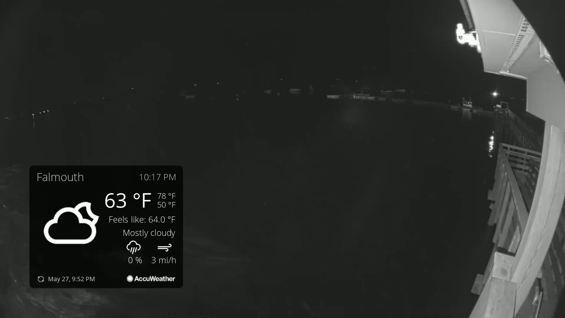 A nighttime, mostly cloudy scene shows a dark body of water with faint lights in the distance, viewed from a wooden deck under a 63°F temperature.