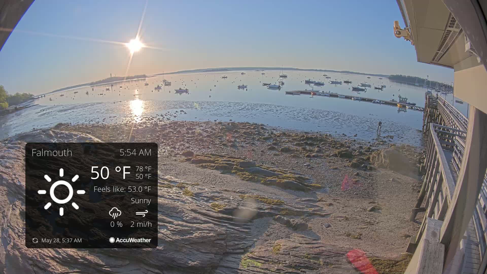 A sunny morning at a harbor shows a calm body of water with many boats at anchor, a rocky shoreline, and a wooden pier in the foreground.