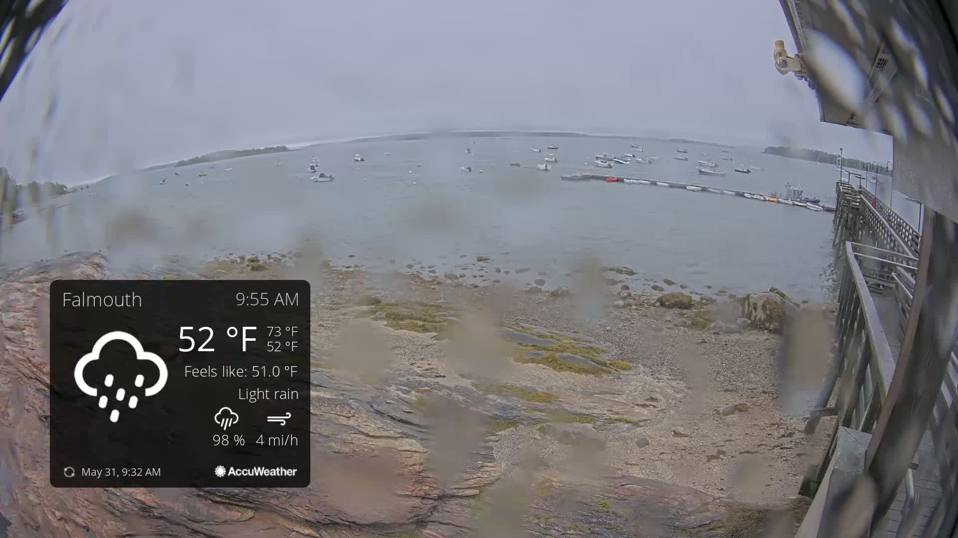 A rocky shoreline with several boats in the water is seen under a light rain with overcast skies.