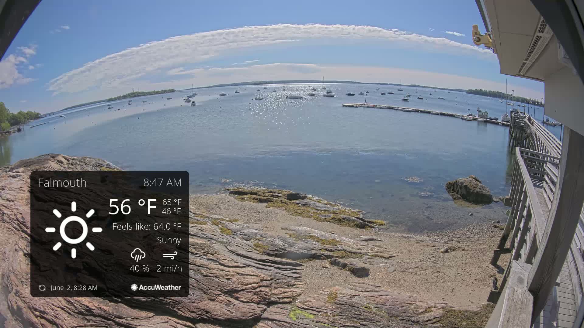 A sunny, partly cloudy day overlooks a calm bay with numerous boats and a rocky shoreline featuring a wooden pier.