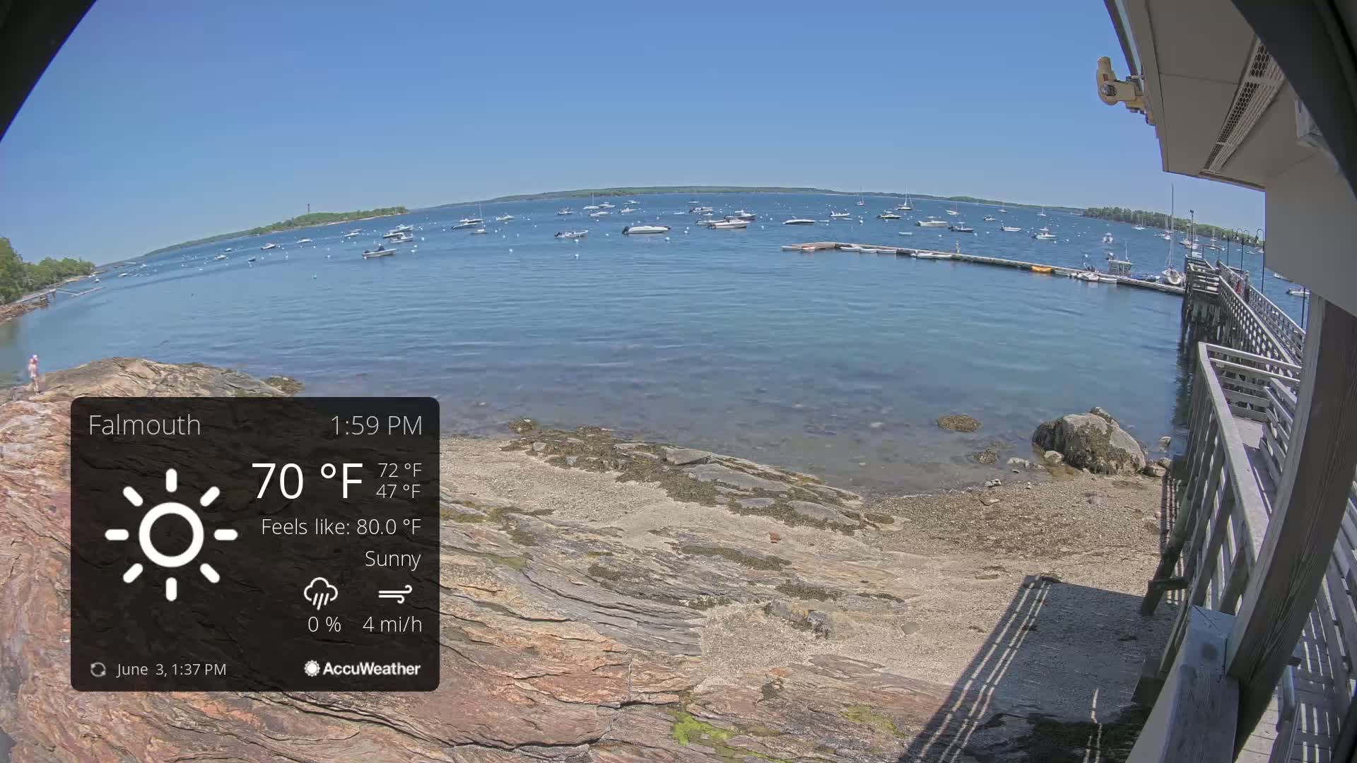 A sunny, calm day overlooking a body of water with numerous boats, a rocky shoreline, and a wooden pier.