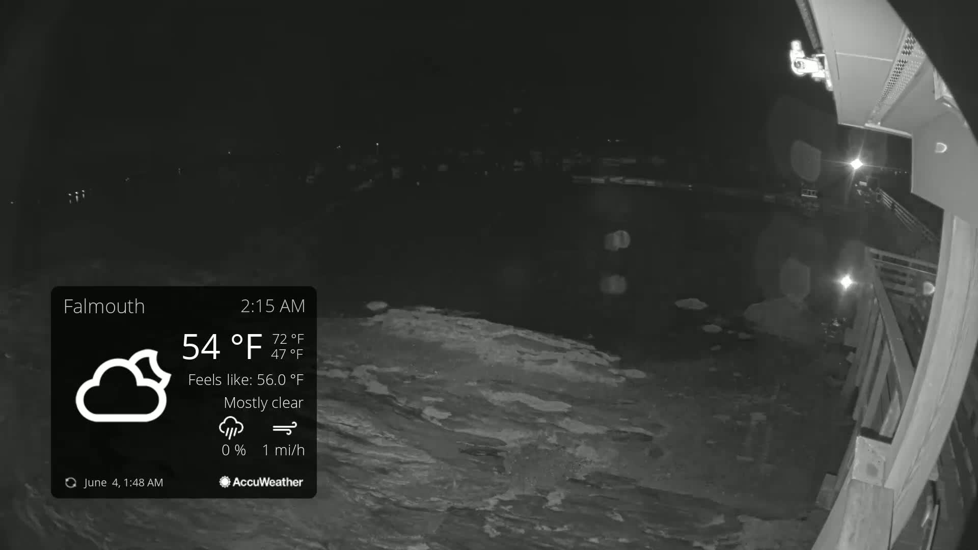 A nighttime, mostly clear view of a dark body of water, possibly a harbor, with a wooden structure and faint lights visible in the distance, under a 54°F temperature.