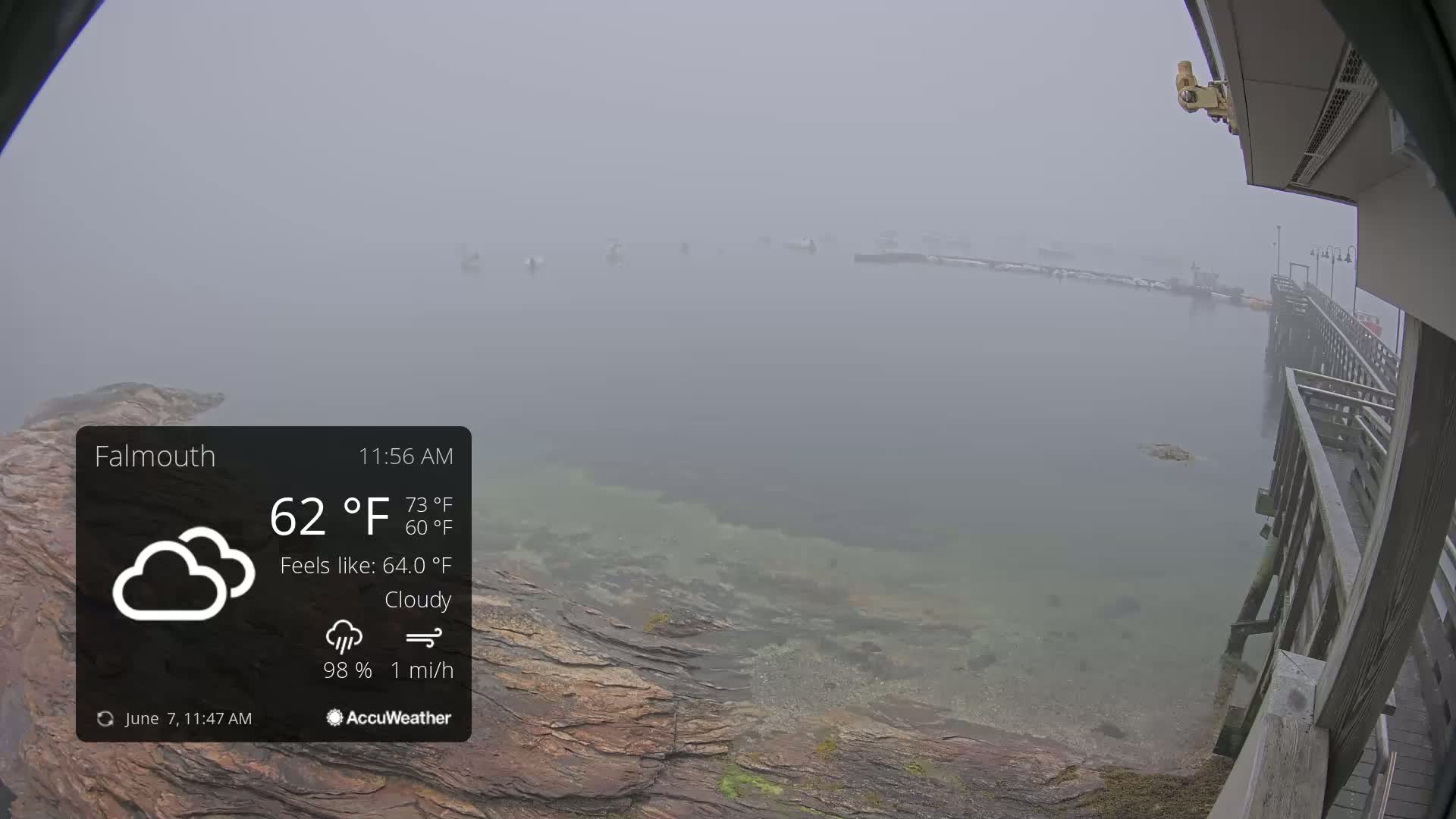 A foggy, cloudy waterfront scene shows a calm body of water with a wooden pier, rocky shoreline, and a few distant boats.