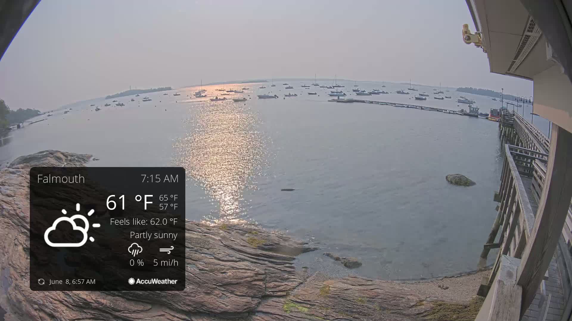 A partly sunny morning view from a wooden pier shows calm water with numerous boats moored in the distance, rocky shoreline nearby, and a slight haze in the air.