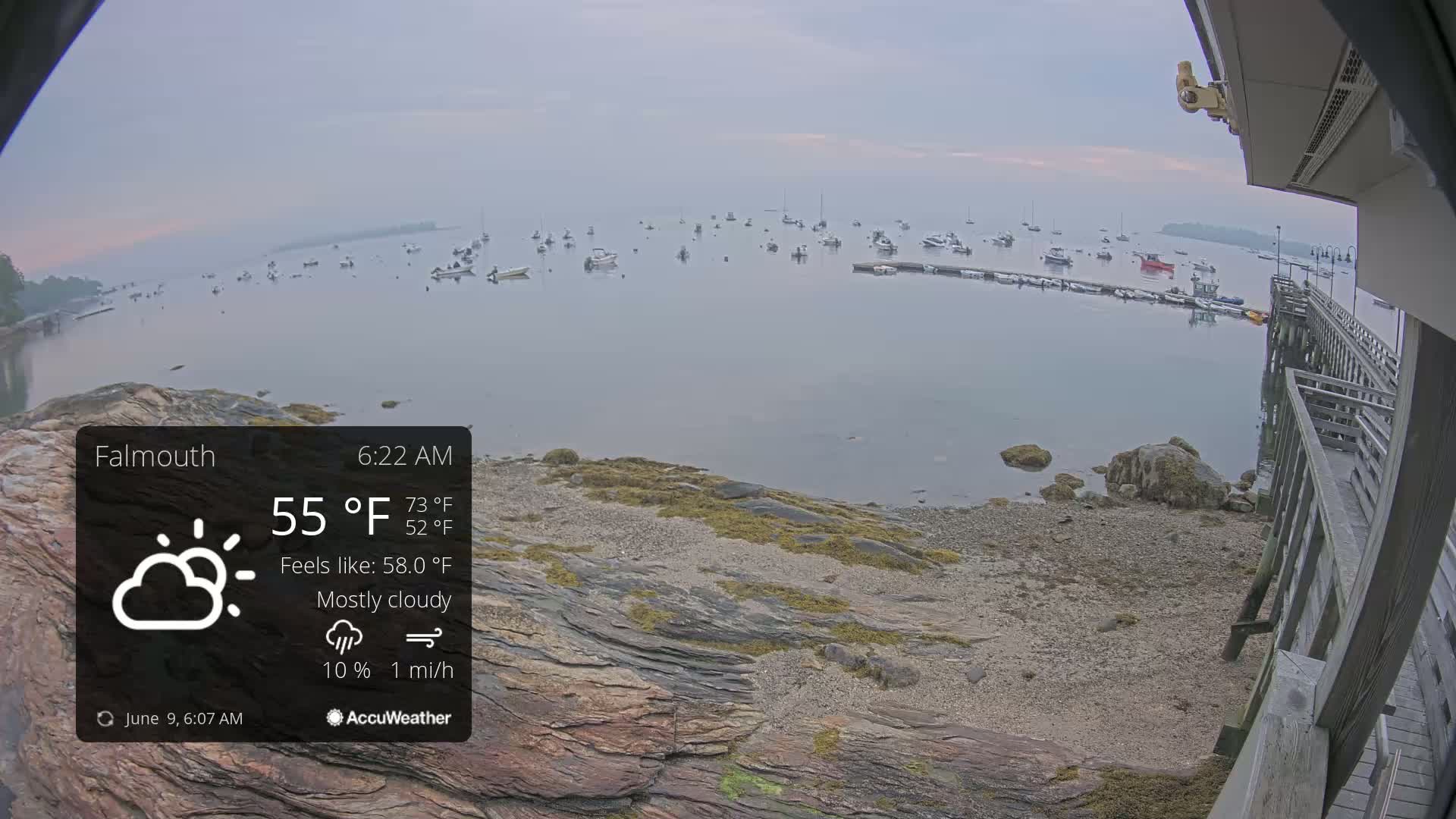 A mostly cloudy morning scene shows a calm body of water with numerous boats, a rocky shoreline, and a wooden pier extending into the water.