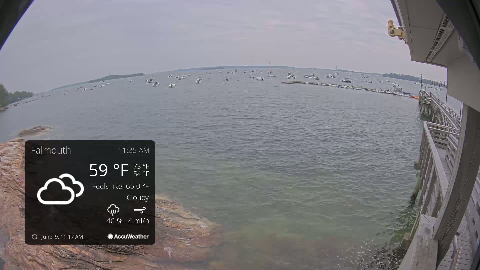 A cloudy, calm day shows numerous boats moored in a bay, viewed from a wooden pier.