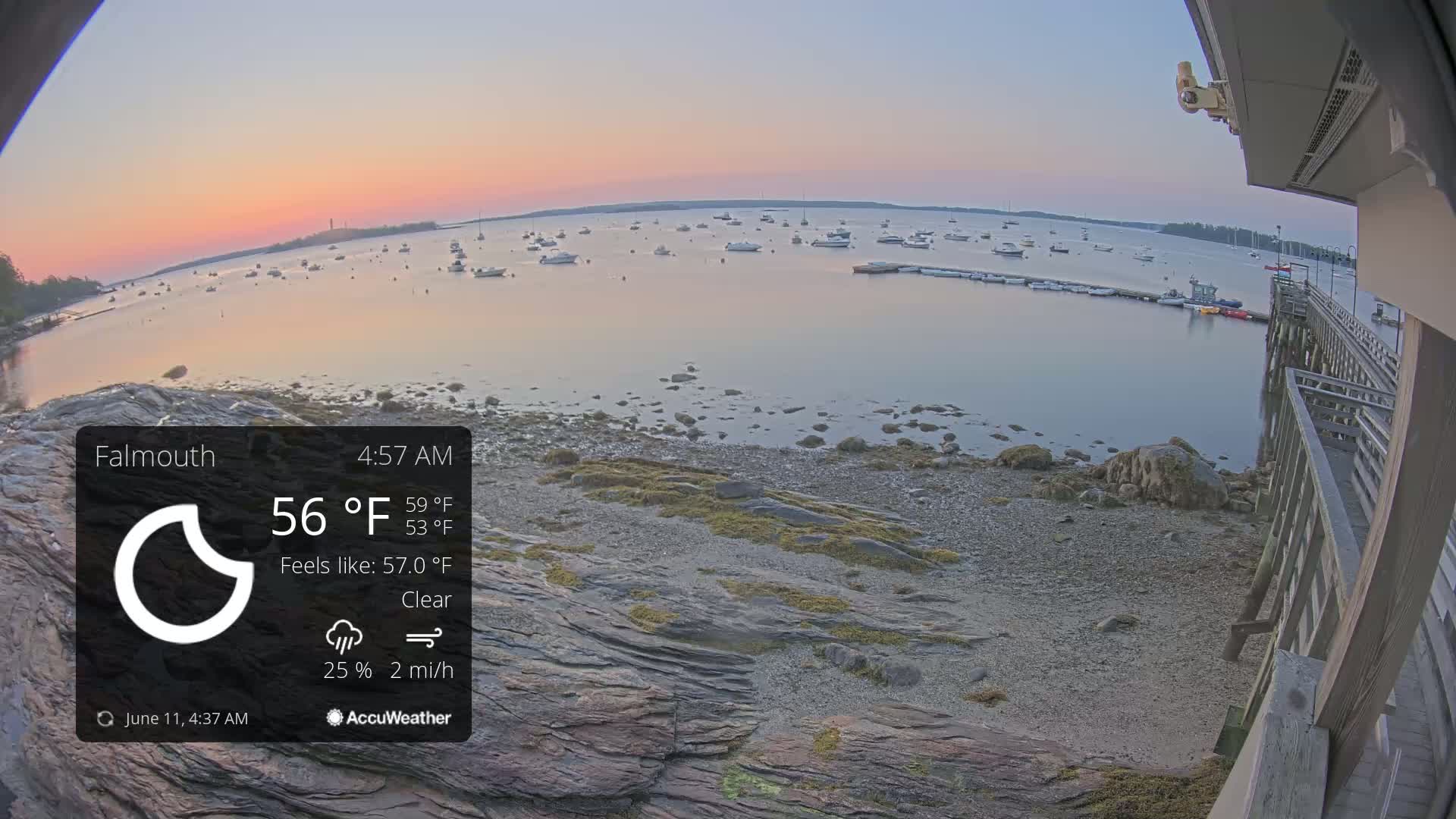 A calm, clear sunrise illuminates a rocky shoreline with numerous boats at anchor in a bay.