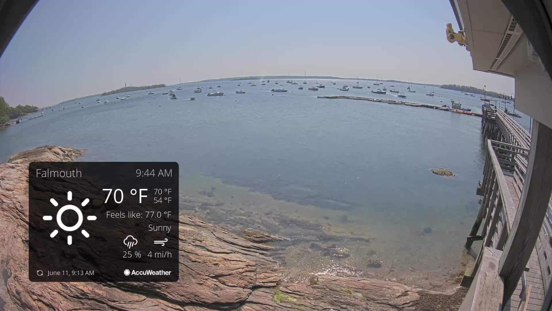 A sunny, calm body of water with numerous boats is viewed from a rocky shoreline with a wooden pier to the right.