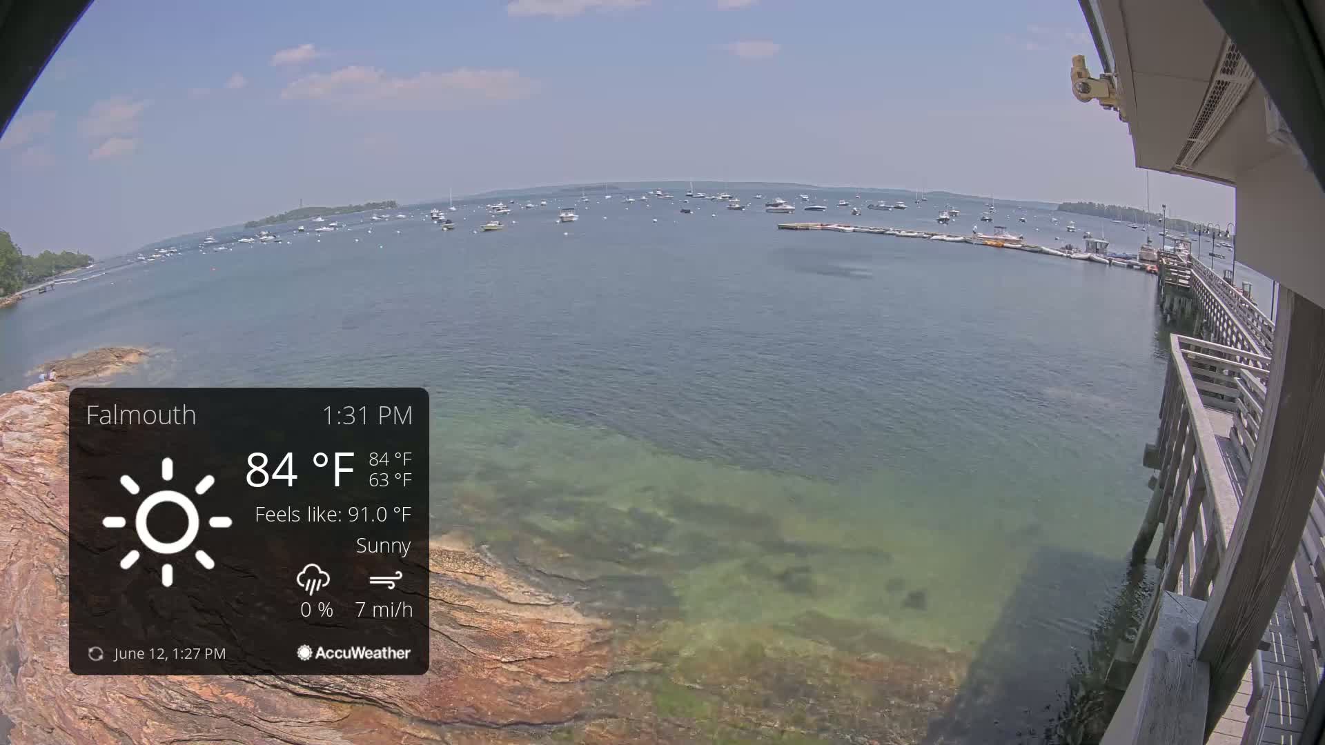 A sunny, calm day with a light breeze shows a body of water with numerous boats, a rocky shoreline, and a wooden pier.