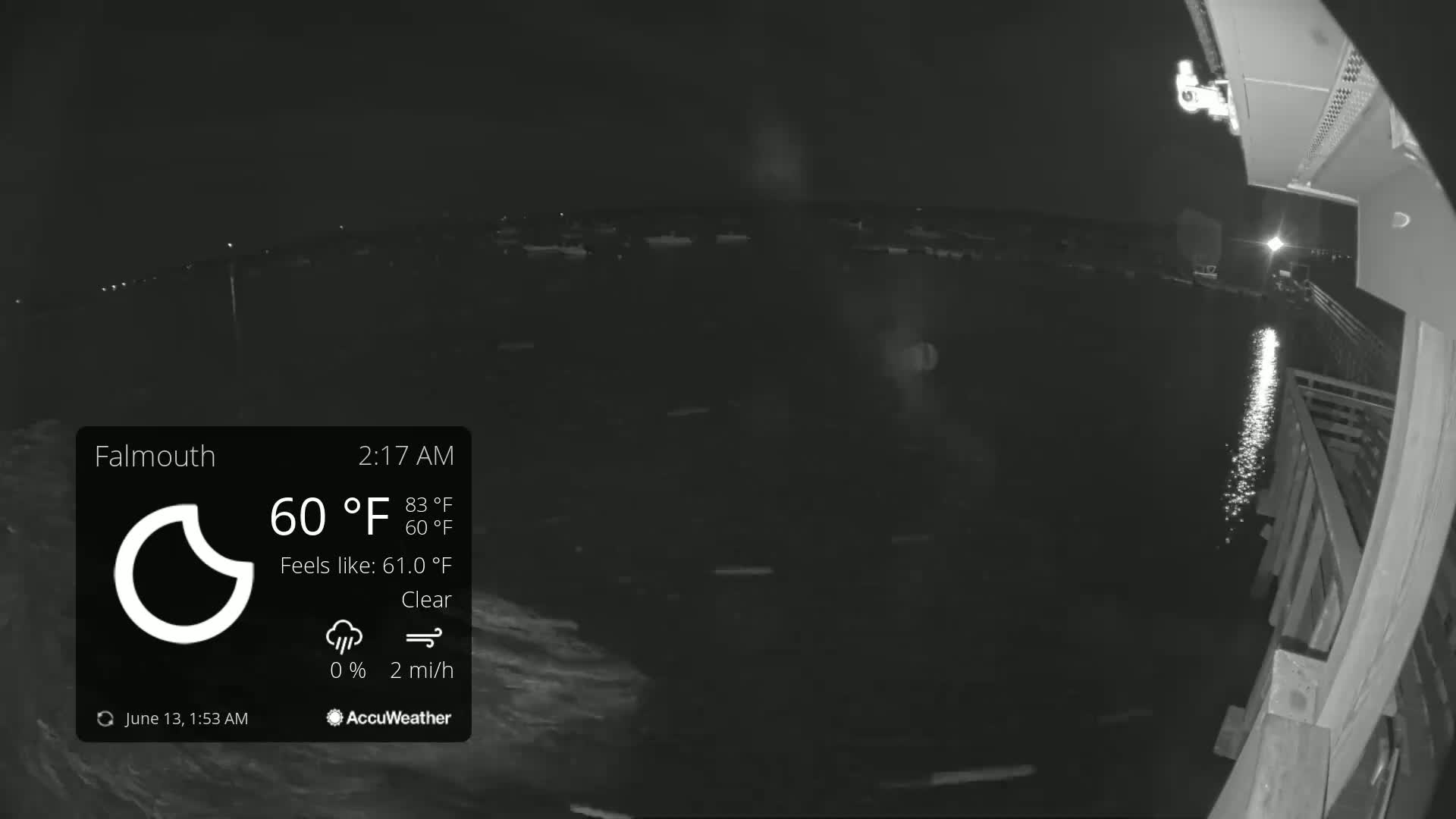 A nighttime view of calm water reflecting lights from a nearby shore, under a clear sky.