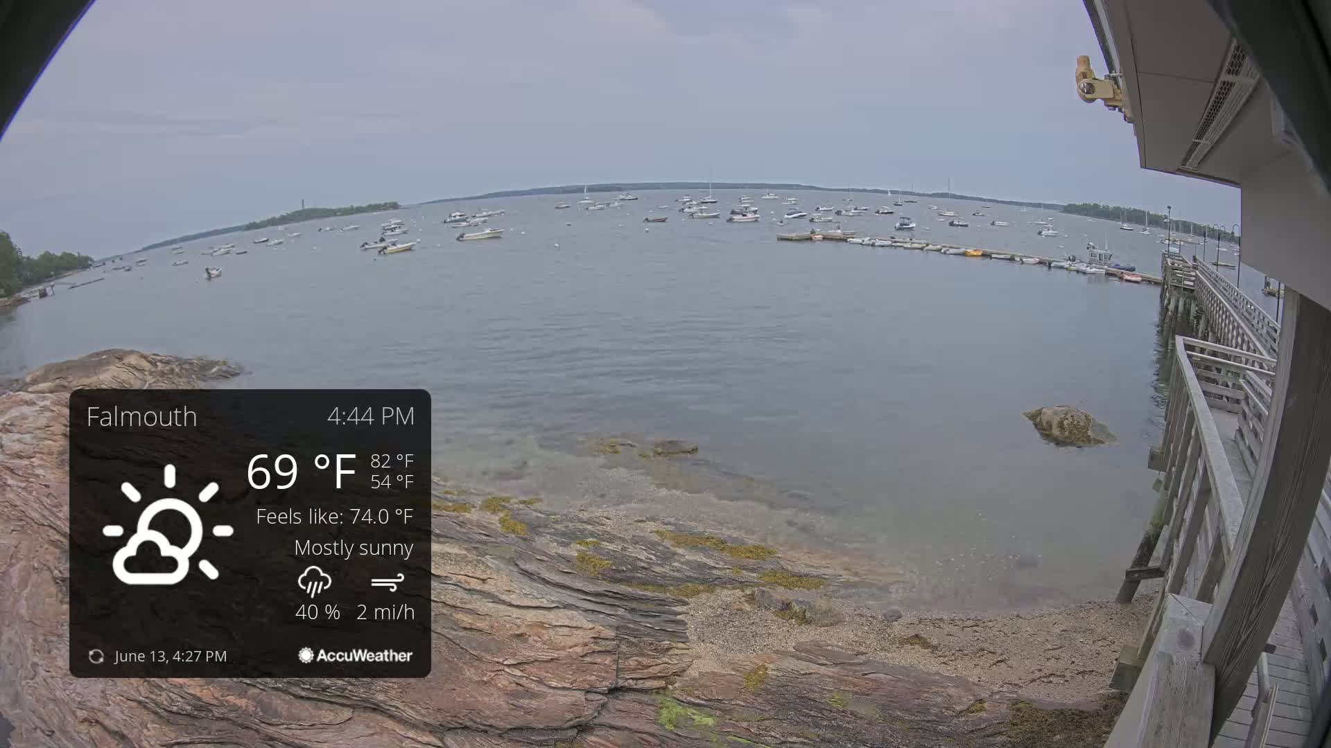 A mostly sunny, calm day shows a rocky shoreline with a wooden pier and many boats in a calm bay.